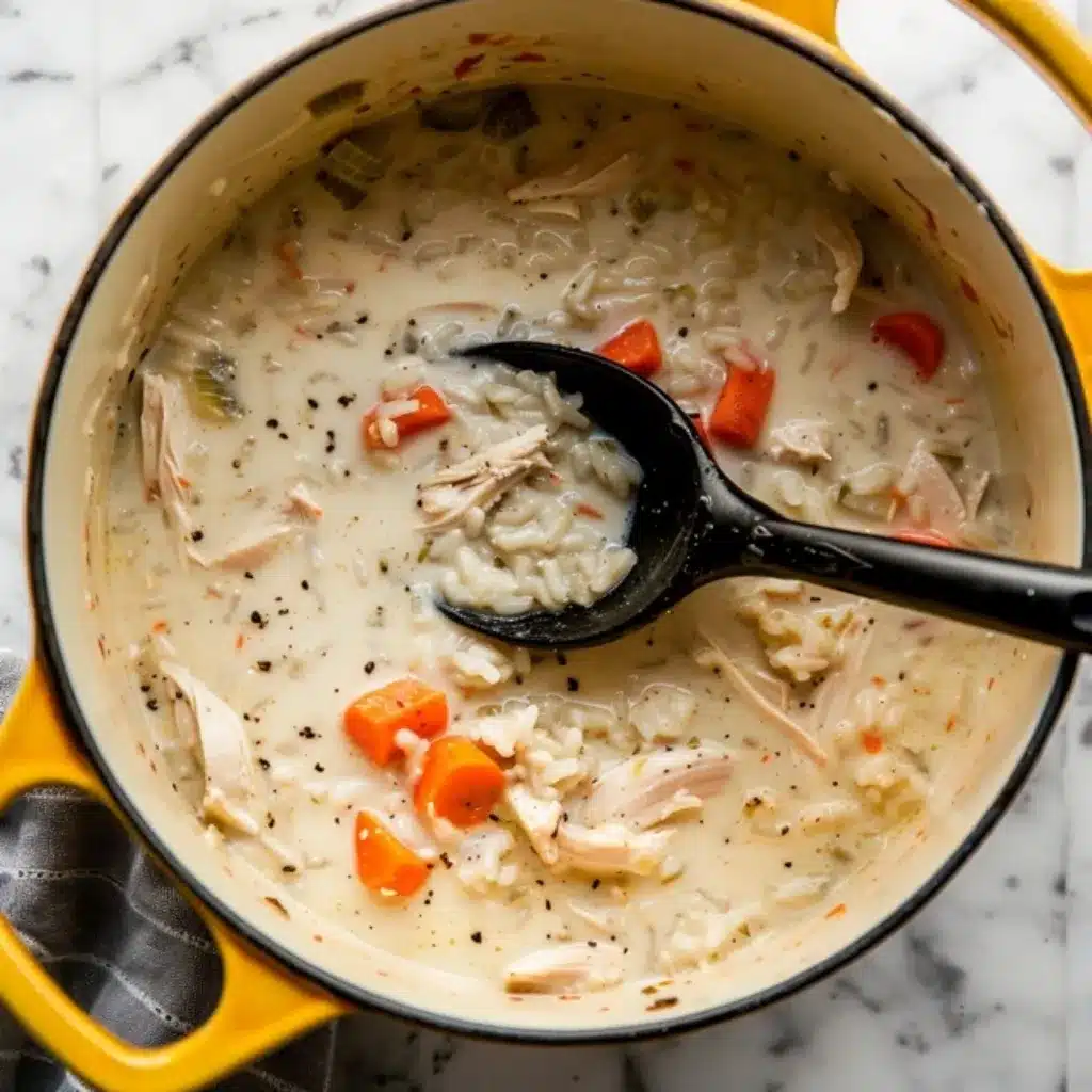 A yellow Dutch oven filled with creamy chicken and rice soup, featuring shredded chicken, carrots, celery, and black pepper, with a black ladle.