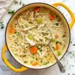 A pot of creamy chicken and rice soup with shredded chicken, carrots, peas, celery, and fresh herbs, served in a yellow Dutch oven with a silver ladle.