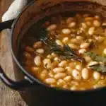 Hearty Tuscan White Bean Soup Recipe 11 A pot of rustic Italian white bean stew with rosemary sprigs and bay leaves, simmering in a golden broth inside a cast iron pot on a wooden table